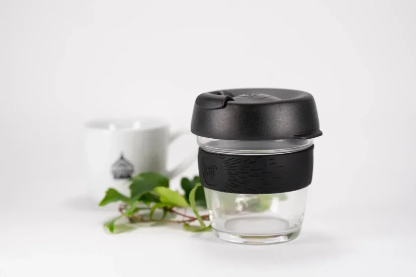 Glass thermal mug with a black lid and black rubber holder, 227 ml capacity, featuring a cup of coffee and a small branch.