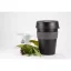 Gobelet thermique noir en plastique avec poignée grise sur une table blanche avec une plante, tasse blanche sur fond blanc.