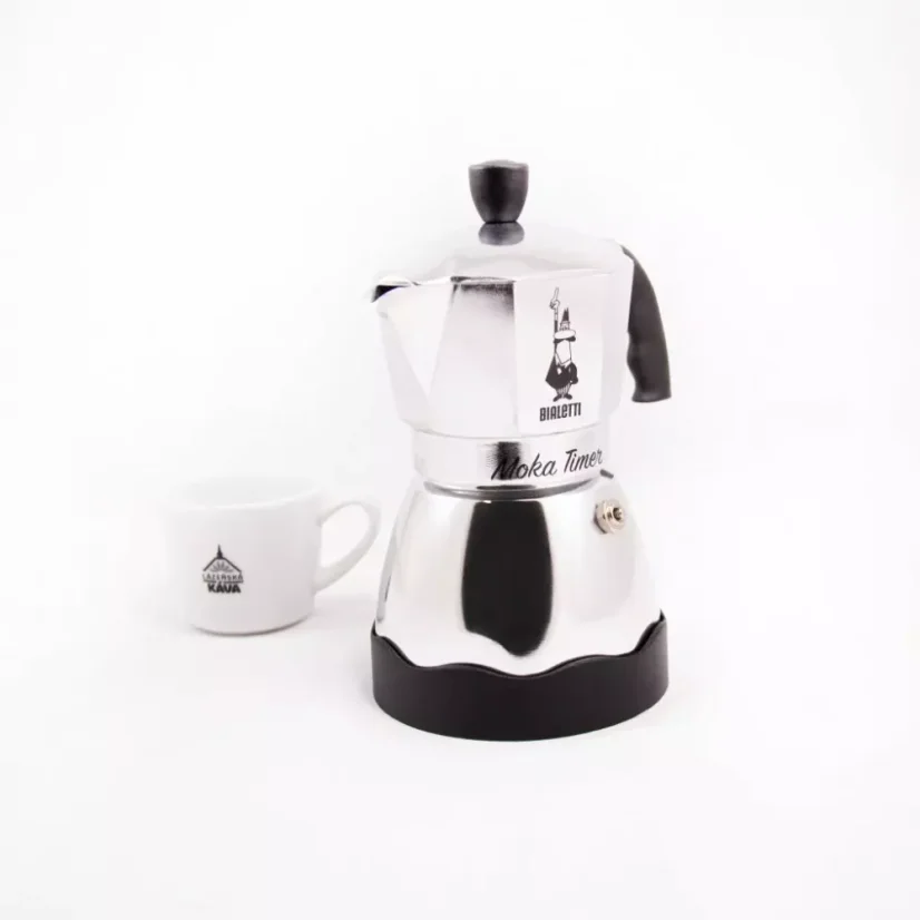 Predný pohľad na striebornú moka kanvicu značky Bialetti Moka Timer a v pozadí biely šálka s motívmi.