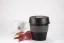 Plastic dark-colored thermal mug with a gray holder on a white table with a red plant and a white mug with a logo on a white background.