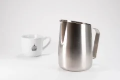 Stainless steel milk frothing jug on a white table together with a porcelain cup.