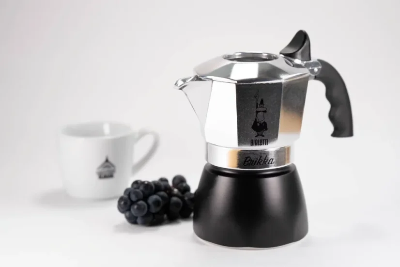 Silver Bialetti Brikka moka pot with a black water chamber, placed on a table with grape wine and a porcelain cup with a coffee logo.