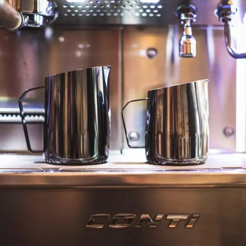 Dve veľkosti nerezových kanvíc na šľahanie mlieka značky Barista a Co Dial in Milk Pitcherb v tmavom prevedení s kávovarom v pozadí.