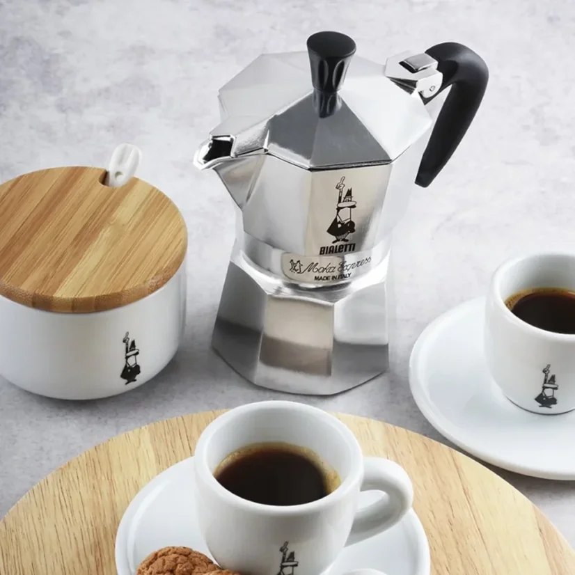 Zilveren mokapot voor 3 kopjes van Bialetti Moka Express op een witte tafel met bereide koffie in porseleinen kopjes en een witte suikerpot met een houten deksel.