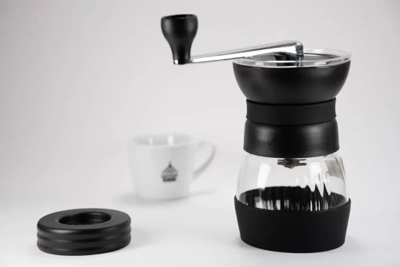 Manual Hario Skerton Pro black coffee grinder on a white table, with a white porcelain cup in the background.