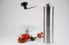 Strieborný nerezový ručný mlynček na bielom pozadí so šálkou kávy a vetvičkou.