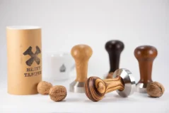 Set of Heavy Temper tampers with wooden handles in various colors on a table with original packaging and a cup with a logo.