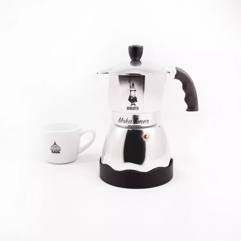 Pohľad zo strany na striebornú moka kanvicu značky Bialetti Moka Timer a v pozadí položený šálka s logom.