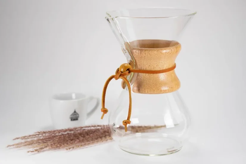 Glass Chemex with wooden handle and leather strap, white coffee cup, and scattered coffee beans on a white table.