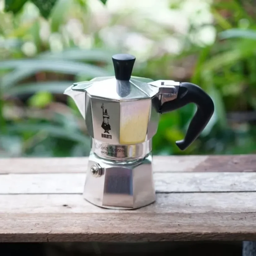 Cafetière Moka Bialetti Express posée sur un banc en bois en pleine nature.
