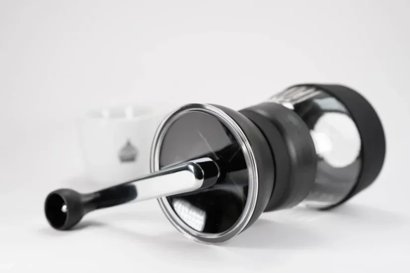 Plastic hand grinder by Hario Skerton Pro lying on a white table next to a porcelain cup with coffee-themed patterns