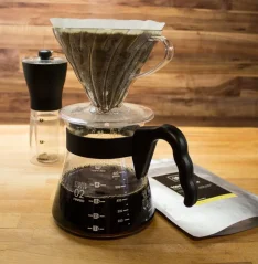 Dripper in plastica, caraffa in vetro con manico, macinacaffè, confezione di caffè su sfondo rustico.