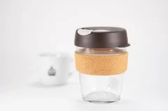 Glass thermal mug with a brown lid and cork holder on a white table, white mug with logo and light background.