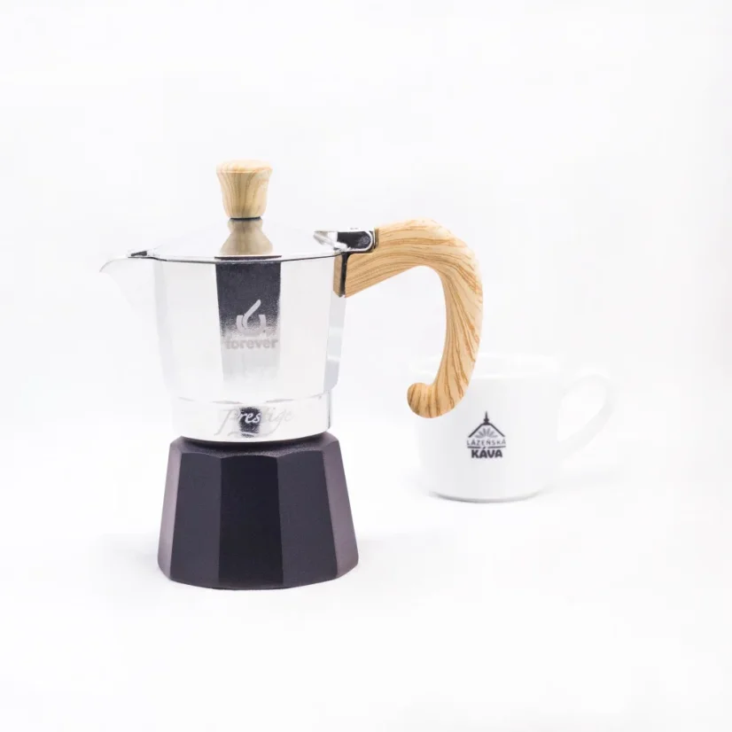 Moka pot with a wooden handle and a black water spout from Forever Miss Moka Woody for 2 servings of coffee with a cup of coffee in the background.