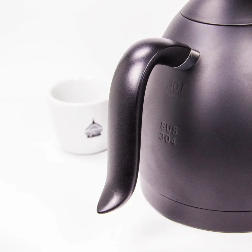 Részletes kép a fekete színű Brewista kanna fogantyújáról, háttérben a fehér kávés csészénkkel.