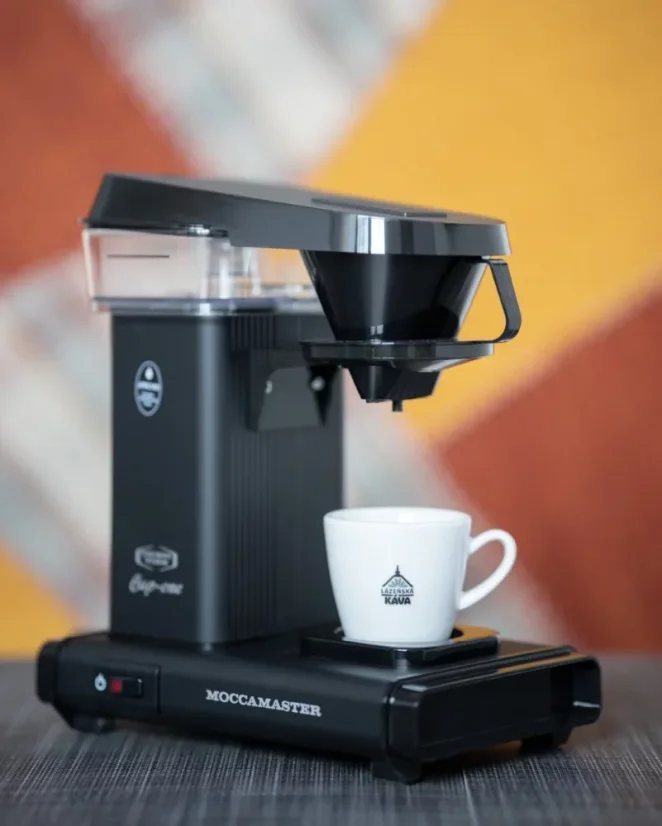 Moccamaster noir Cup One sur une table grise avec une tasse blanche sur un fond coloré.