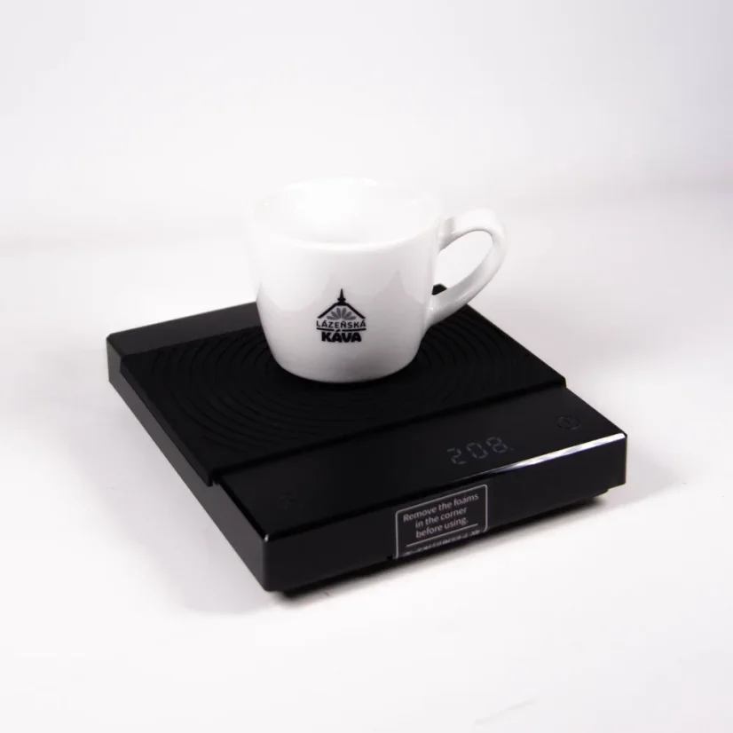 Čierna baristická digitálna váha Black Mirror s čiernou gumovou podložkou a na nej položený šálka kávy.