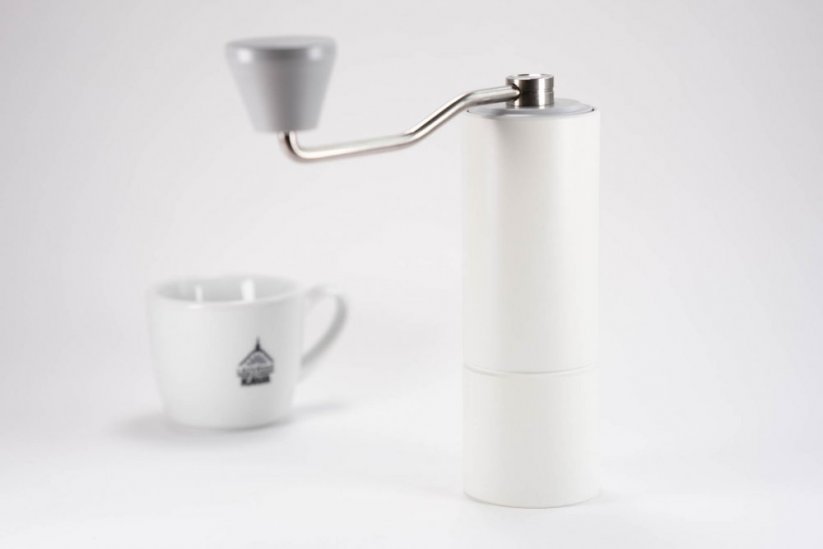 Timemore C2 kézi kávédaráló fehér színben, szürke fogantyúval. Rozsdamentes acél csiszolókövekkel rendelkezik. A háttérben egy csésze a Spa Coffee logójával.
