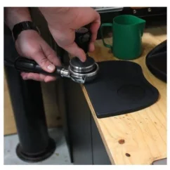 Barista qui tasse le café dans le porte-filtre sur un tapis en caoutchouc noir de Rhinowares posé sur une table en bois.